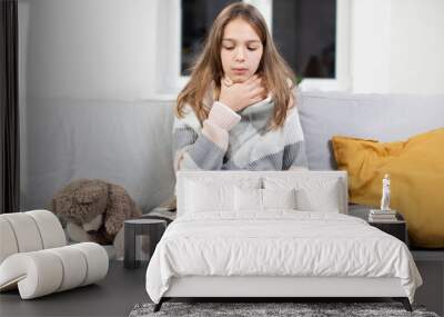 Pharyngitis, sore throat concept. Sick Caucaisna teenage girl touching sore throat, feeling unwell, while sitting on the sofa at home, covered with blanket and drinking hot tea Wall mural