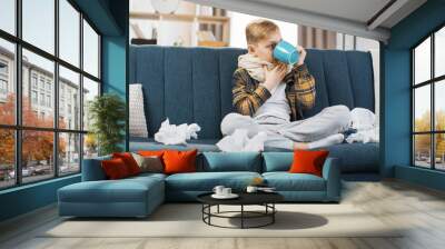 Pharyngitis, sore throat concept. Sick caucasian teenage boy touching sore throat, feeling unwell, while sitting on the sofa at home, and drinking hot tea. Wall mural