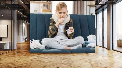 Pleasant tired sick teen boy with scarf around neck, sitting on soft couch and coughing, suffering from flu or cold and sore throat. Flu treatment at home. Wall mural