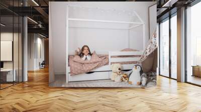 Portrait of a beautiful caucasian little girl smiling while relaxing in her bedroom during a leisure day. Cheerful female kid lying on scandinavian wooden house frame bed. Wall mural