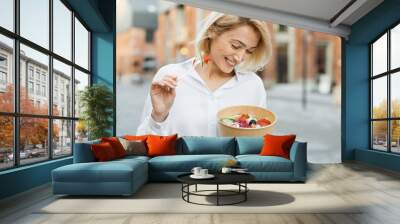 Portrait of a young attractive woman eating salad at the background of buildings of city. Beautiful young female enjoying a fresh salad outdoor. Cute blond girl eating a healthy meal. Wall mural