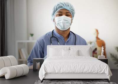 Portrait of african american male in blue scrubs wearing face mask, medical cap and latex gloves holding stethoscope around neck in workplace. Health professional getting ready for examination. Wall mural