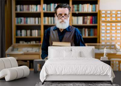 Portrait of attractive bearded old man wearing shirt and leather vest, high school teacher or librarian, holding books in hands, standing in vintage library interior. Library worker, happy book day Wall mural