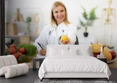 Portrait of smiling female nutritionist doctor sitting at table with vegetables and fruits in clinic. Positive mature Caucasian female weighing lemon while writing healthy food recipe on clipboard. Wall mural