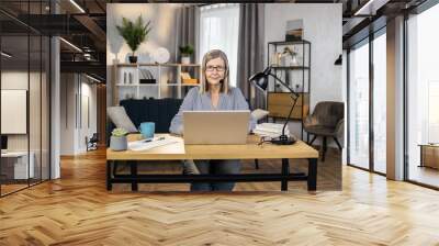 Portrait of smiling female worker in hands-free headset sitting behind laptop at office desk in spacious workplace at home. Mindful remote employee scheduling group meeting with teammates online. Wall mural