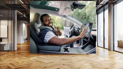 Portrait of young smiling handsome African man with beard sitting inside his modern self-steering car and wiping dust from touchscreen display with microfiber cloth, looking at camera Wall mural