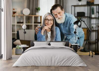 Positive smiling senior wife and husband coworkers in casual wear posing at bright home with modern gadgets on table. Concept of cooperation, people and technology, online work at home in retirement Wall mural