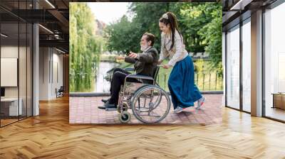 Pretty hipster young woman with dreadlock hair pushing wheelchair with elderly man, having fun in the city park in front of lake. Handicapped man is happy during walk with his granddaughter. Wall mural