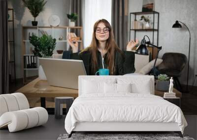 Pretty woman freelancer in eyeglasses sitting at table with closed eyes and relieving stress by meditation at workplace. Concept of relaxation and harmony, no stress free relief at work Wall mural