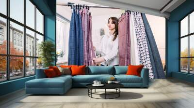 Pretty young Caucasian female designer or tailor standing near clothes rack with stylish pants, working in her atelier studio Wall mural