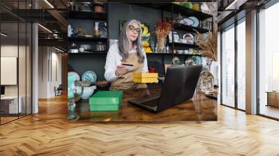 Professional aged woman holding discount card and yellow box while sitting at decor store. Saleswoman using modern laptop at workplace. Wall mural