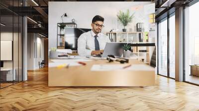 Professional Arab Muslim male architect using laptop computer, sitting on the desk while working on construction plan, using sketches at his workplace. Wall mural