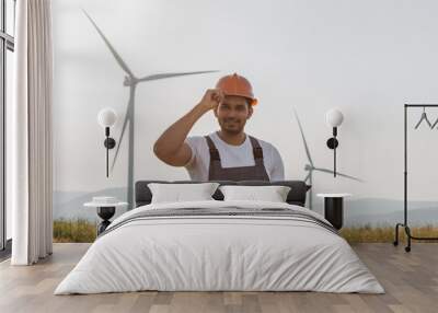 Professional technician in helmet and overalls smiling oj camera while standing on eco farm with wind turbines. Indian man using digital tablet while controlling process of clean energy making. Wall mural