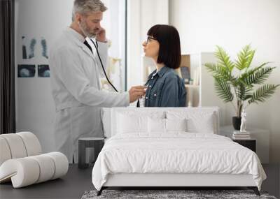 Side view of focused man in white coat using stethoscope while beautiful female in eyeglasses sitting quietly on couch. Professional specialist hearing heart's rate and rhythm in examination room. Wall mural