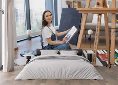 Side view of talented artist sitting on floor in lotus position with canvas on knees and drawing with brush. Beautiful lady with long brown hair painting portrait of man at comfortable home studio. Wall mural