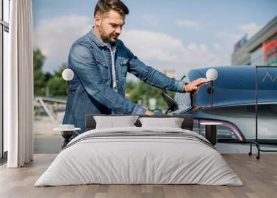 Side view of young handsome bearded man in jeans shirt, plugging wire into the car socket to charge his new luxury modern electric car at outdoor charging station in the city Wall mural