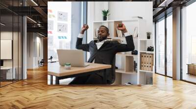 Sleepy and tired african american man in formal wear yawning and stretching arms after hard working hours on wireless laptop at office. Concept of people, technology and overwork. Wall mural