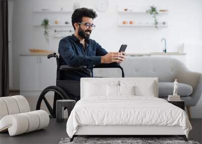 Smiling arabian adult in wheelchair looking at cell phone screen while having rest in comfortable dining room. Cheerful positive man checking social media using internet connection in apartment. Wall mural