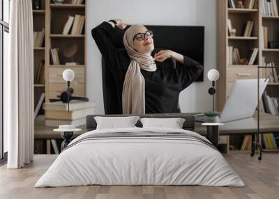 Smiling muslim woman relaxing by stretching her body while sitting in front of pc computer laptop at the wooden working table over comfortable office. Wall mural