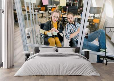 Smiling young disabled hipster businesswoman with blond dreadlocks, sitting in wheelchair, reading sticky notes on a glass wall and writing her ideas while brainstorming with male coworker in office Wall mural