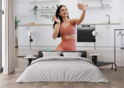 Smiling young woman with dark hair having video call on modern smartphone during domestic workout. Sporty female waving hand on mobile camera while sitting on mat with dumbbells and bottle of water. Wall mural