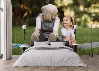 Sport, gymnastics, fitness and physical activity concept, where active smiling Muslim mother having fun with her little daughter, doing stretching exercises on yoga mat outdoors in the park Wall mural