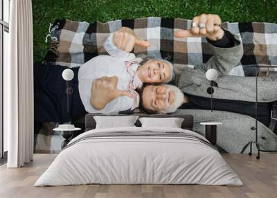 Stylish senior couple smiling and showing thumbs up while lying on soft checkered blanket. Happy aged man and woman spending time together on fresh air. Wall mural