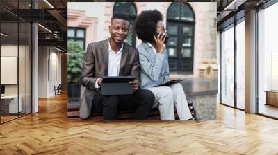 Successful afro businessman smiling and looking at camera with digital tablet in hands while his female colleague sitting near and talking on mobile. Two people working outdoors. Wall mural