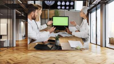 Teamwork of four multiethnic doctors wearing white lab coats and pointing on laptop with green screen while working in polyclinic boardroom. Concept of effective team cooperation in healthcare field. Wall mural