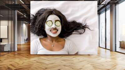 Top-view of smiling African woman making spa procedures with mud facial mask and fresh cucumber slices on eyes, laying on bed. Wellness and skincare concept Wall mural