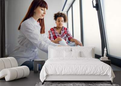 Trauma on the child's leg. Female Caucasian doctor orthopedist, examining foot of pretty mixed race teen girl in clinic. Child suffering from pain at doctor's office Wall mural