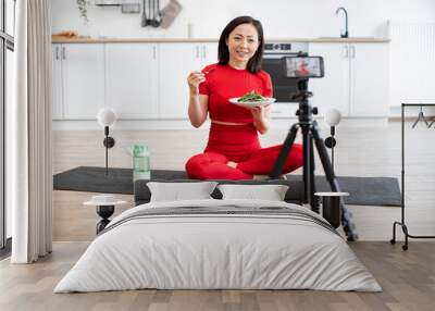 Woman in red activewear sitting on yoga mat eating salad while filming fitness video in modern kitchen. Healthy lifestyle, nutrition, and social media influencer concept. Wall mural