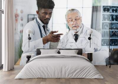 Young african american and senior caucasian doctors in white lab coats standing near flip chart with laptop and analysing x rays scan. Two medical specialists having consultation indoors. Wall mural