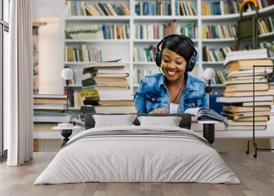 Young african smiling woman in a good mood listening to music while studying in a library. Education concept Wall mural