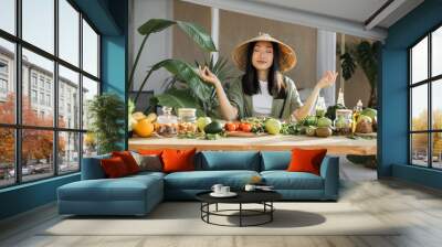 Young asian woman sitting near table with organic vegetables in yoga position while making salad, enjoying healthy diet, indoor at light exotic kitchen studio interior. Weight loss concept. Wall mural