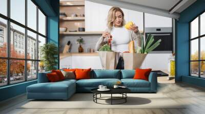 Young blond woman holding grocery shopping bag with vegetables standing in the modern light kitchen at home. Attractive female unpacking paper bag with fresh products. Wall mural