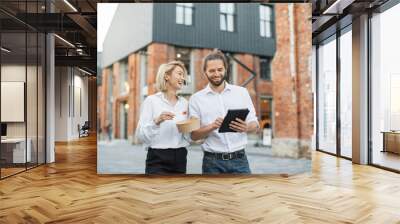 Young business colleagues in formal clothes walking on street and discussing some working issues during break. Young man using tablet and woman eating healthy salad. Wall mural