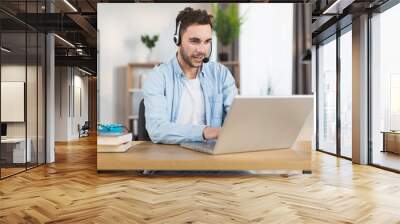 Young caucasian man in headset sitting at desk and having video call on modern laptop. Concept of gadgets, remote work and communication. Wall mural