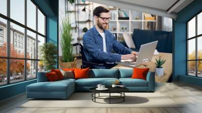 Young man with special needs in casual clothes and eyeglasses working on wireless laptop. Male freelancer working from home while sitting in wheelchair. Wall mural