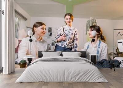 Young smiling fashion designer posing with scissors at working place and looking at camera while two pretty dressmakers sitting at the table and discussing the working process. Wall mural