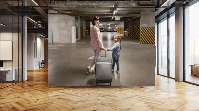 Youthful lady with her child clutching travel suitcase searching for vehicle before starting journey. Happy mom and small daughter, attired in informal wear, stroll through subterranean parking area. Wall mural