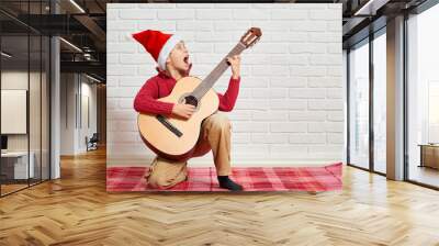 boy playing music on guitar, dressed in a red woolen sweater and santa hat, sitting on a red checkered blanket, white brick wall on background Wall mural