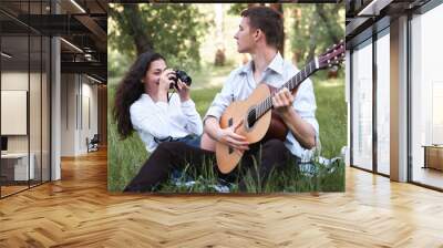 young couple sitting in the forest and playing guitar, taking photo on old camera, summer nature, bright sunlight, shadows and green leaves, romantic feelings Wall mural