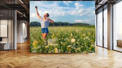 Young woman on the meadow wearing headphones listening music from the smart phone on a sunny summer day Wall mural