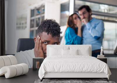 A Sad looking Black man is working on desk and there are two colleagues are gossiping at background behind of him Wall mural