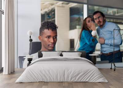 Black man is working on desk and there are two colleagues are gossiping at background behind of him Wall mural