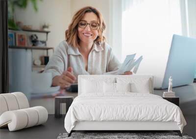 Beautiful middle-aged woman in glasses and paper bills joyfully laughing while calculating revenue grows at home office. Small business, successful investment, bull market, money savings concept image Wall mural