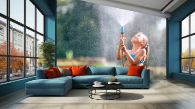 Cute little girl pours herself from the hose, makes a rain. Hot summer day pleasure Wall mural