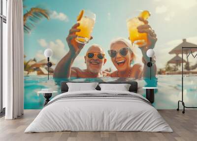 Elderly grey-haired couple cheerful laughing while they rise up alcoholic cocktails with orange juice in swimming pool during summertime exotic vacation together Happiness of senior retirement concept Wall mural