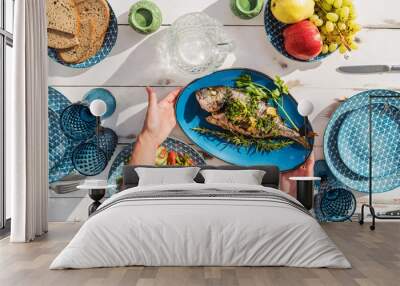 Gilt-head bream also known as Orata grilled on a barbeque grill served by a woman on a white wooden table with vegetable salad and fruits. Healthy seafood preparation concept photo. Wall mural